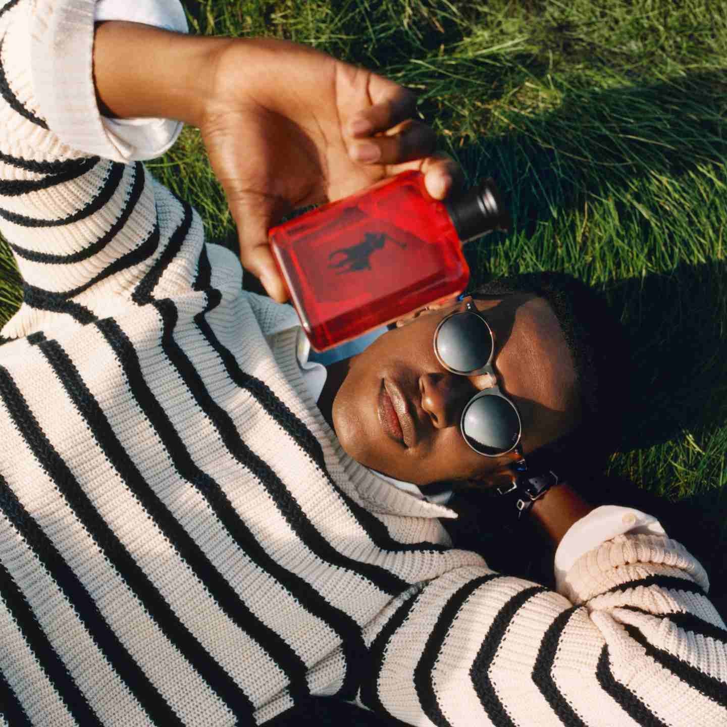 Man lying on grass wears black-and-white striped sweater and holds red fragrance bottle.